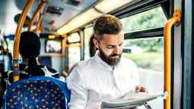 A man is sitting on the bus. He is wearing a white crinkled shirt, and is reading the newspaper. 