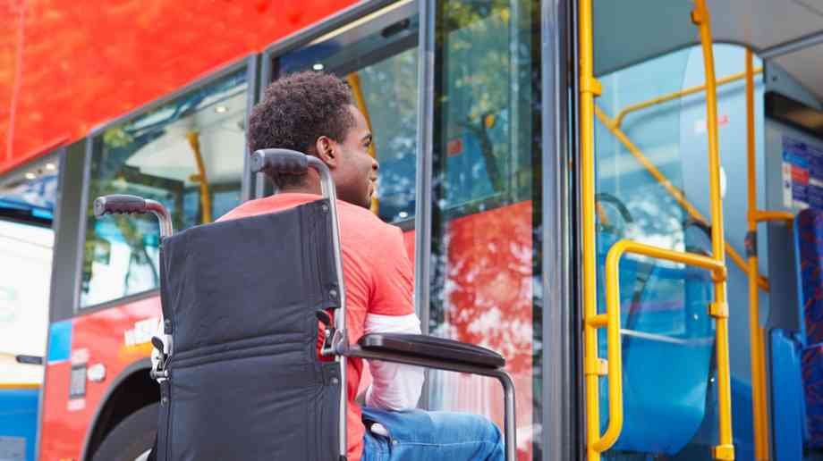 A young man in a wheelchair is sat in front of a red London bus. He is smiling but we can't see all of his face as it is turned away from the viewer. 